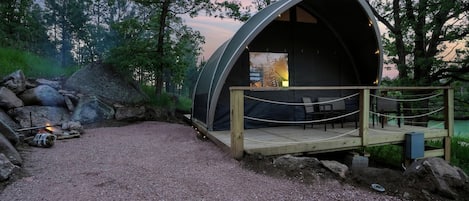 Eine halbkreisförmige, überdachte Außenanlage mit Holzterrasse und Sitzgelegenheit, umgeben von einer natürlichen Landschaft und einem Kiesweg.
