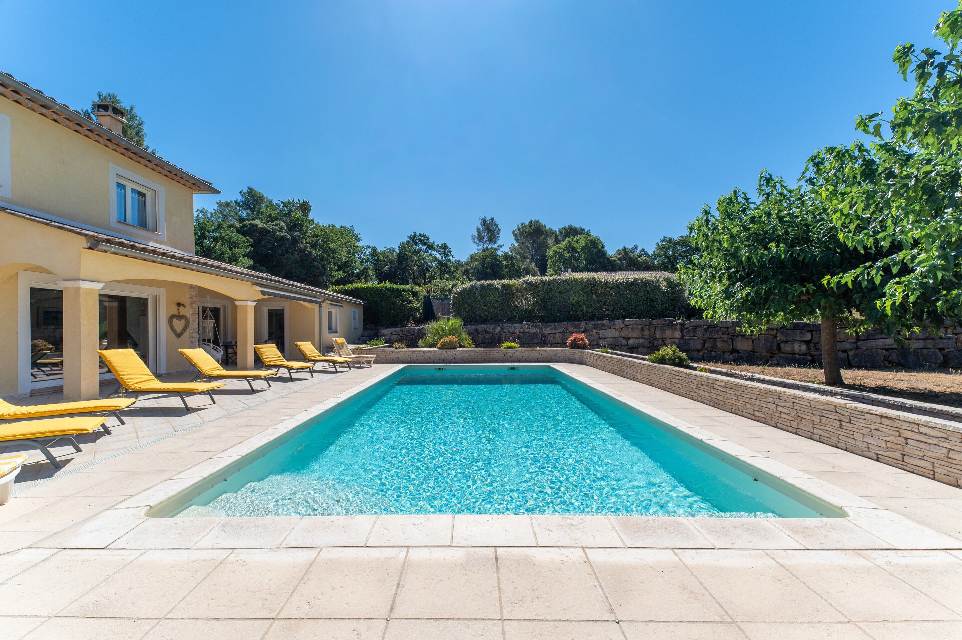 Una piscina con tumbonas, un muro de piedra y un edificio con tejado de baldosas.