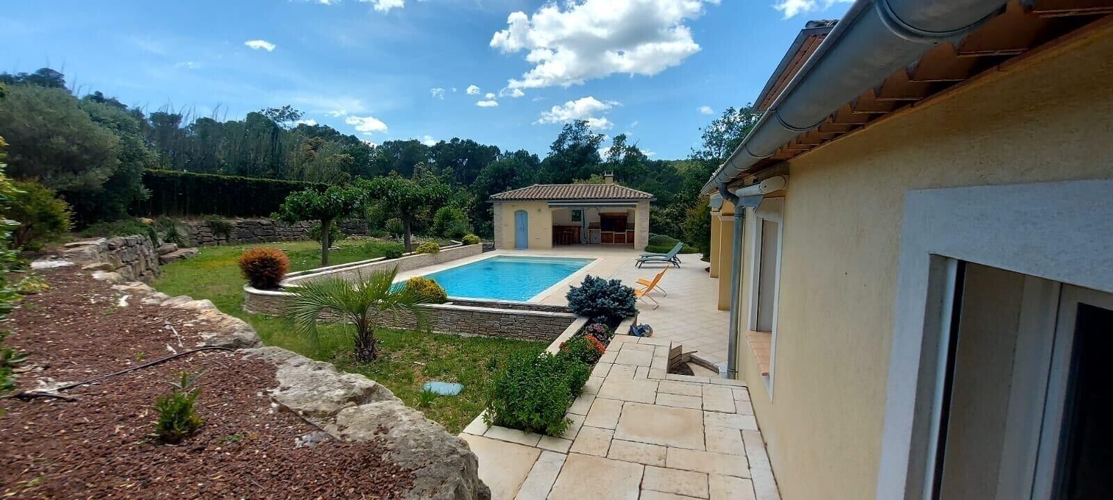 A pool with a stone wall, a small building, and a garden with trees and shrubs.