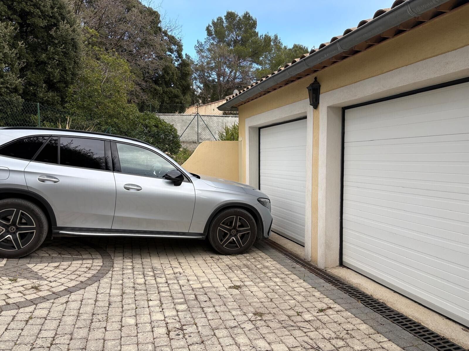 Um SUV prateado estacionado numa entrada de garagem com uma porta de garagem e uma cerca ao fundo.