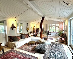 A cozy room with a large sectional sofa, multiple rugs, a standing lamp, and a guitar on the wall. - Charming Rågeleje Summer House with Sauna (Græsted)