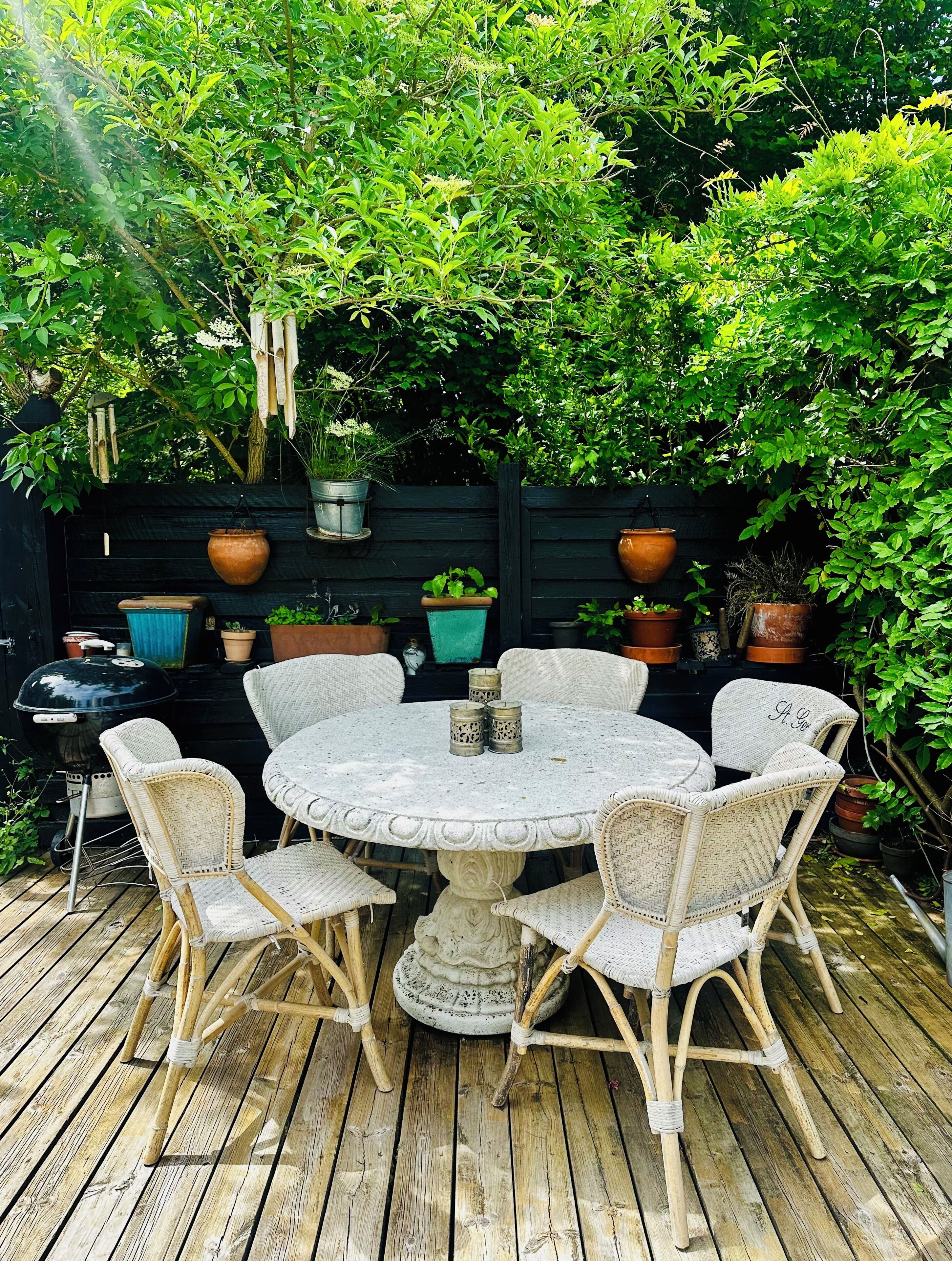 Una mesa redonda al aire libre con sillas de mimbre, rodeada de plantas en macetas y una terraza de madera.