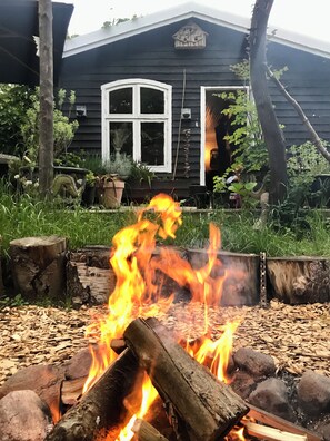 A fire pit with logs in a garden area, surrounded by mulch, with a house in the background. - Charming Rågeleje Summer House with Sauna (Græsted)