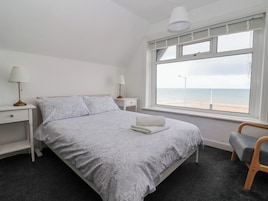 Un dormitorio con cama, dos mesitas de noche, lámpara, una silla y un ventanal con vista a la playa.