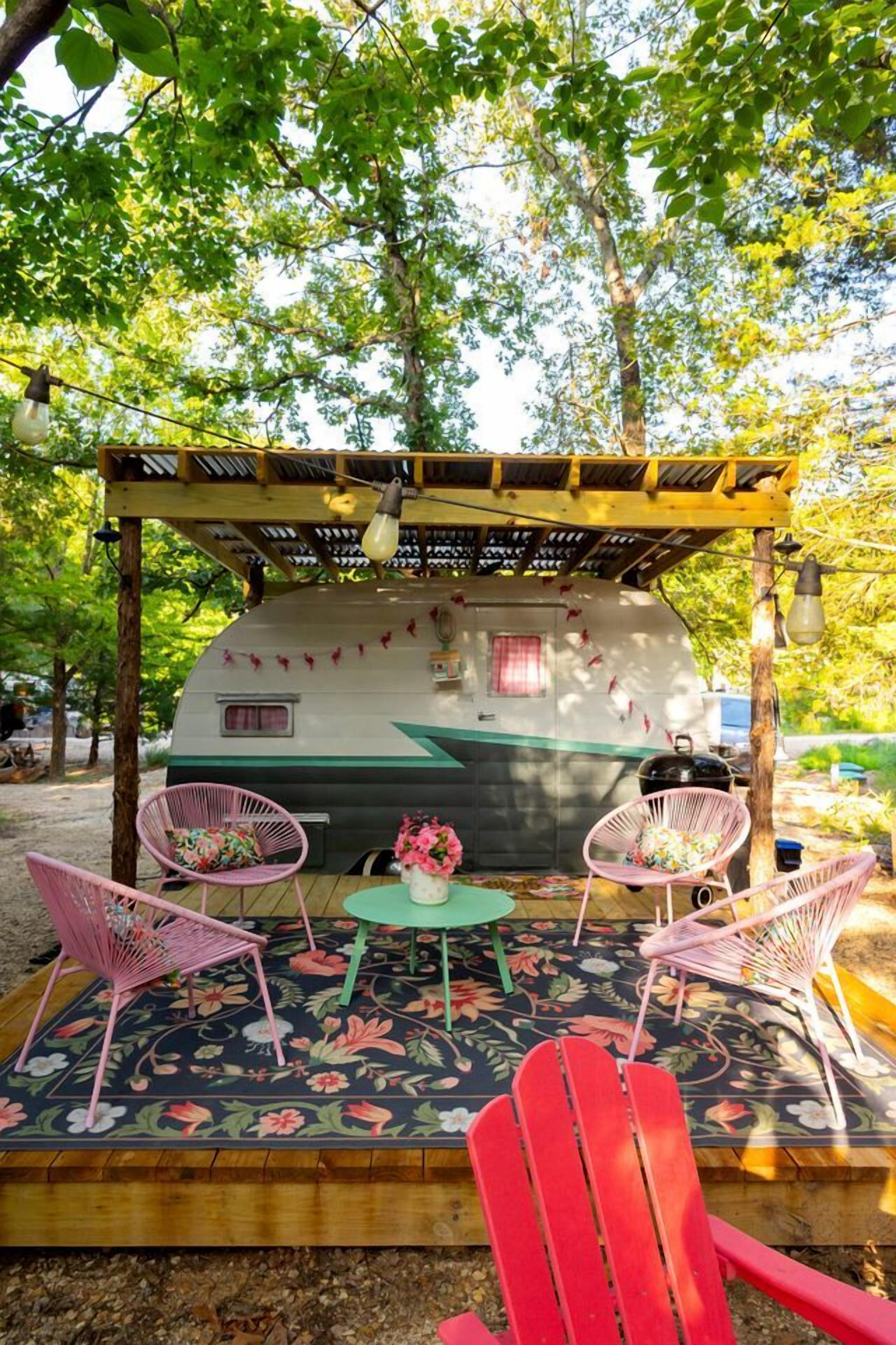 A covered outdoor seating area with a camper, two chairs, a table, and a floral rug.