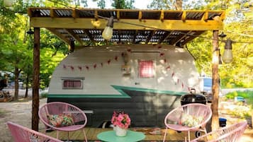 A covered outdoor seating area with a camper, two chairs, a table, and a floral rug.