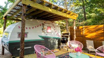 Een oldtimer camper met een overdekte veranda, roze stoelen, een tafel met bloemen en een barbecue.