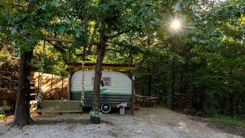 A vintage camper parked in a forested area with a bench and picnic table nearby.