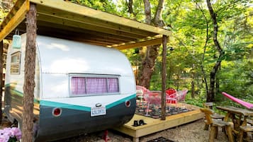 Una caravana antigua estacionada bajo un refugio de madera en un área boscosa, con una mesa y sillas de picnic.
