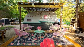 A covered outdoor seating area with a table and chairs, a small table with a vase of flowers, and a covered RV in the background.