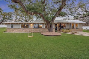 A single-story house with a large tree in front, a well-maintained lawn, and a porch with outdoor seating.