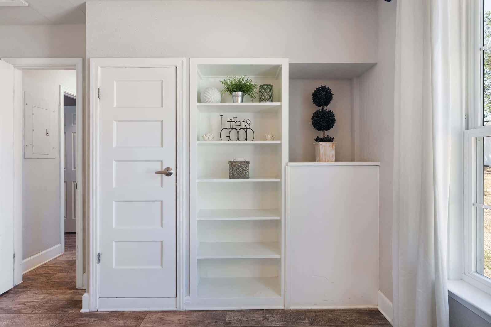 Un espace intérieur aux murs blancs, avec une porte, une bibliothèque intégrée et une fenêtre.