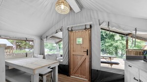 Una carpa con una mesa de madera, asientos tipo banco, una puerta y vistas al exterior.