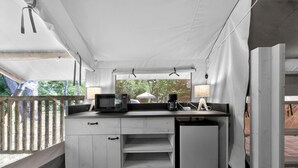 A compact kitchenette with a microwave, coffee maker, and lamp, under a white tented roof with a view of greenery outside.