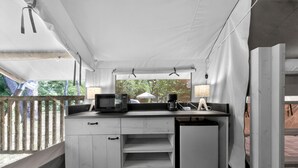 Una kitchenette compacta con microondas, cafetera y lámpara, bajo un techo blanco cubierto, con vistas a la vegetación.