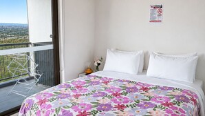 Ein Schlafzimmer mit einem Bett, weißen Kissen, einer blumigen Tagesdecke, einem Nachttisch mit einer Vase und einem Balkon mit Aussicht.
