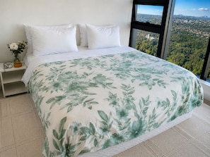 A bed with a floral bedspread, white pillows, and a view of a cityscape through a window. - Cozy apartment Sydney Olympic Park free parking 03 (Sydney Olympic Park)