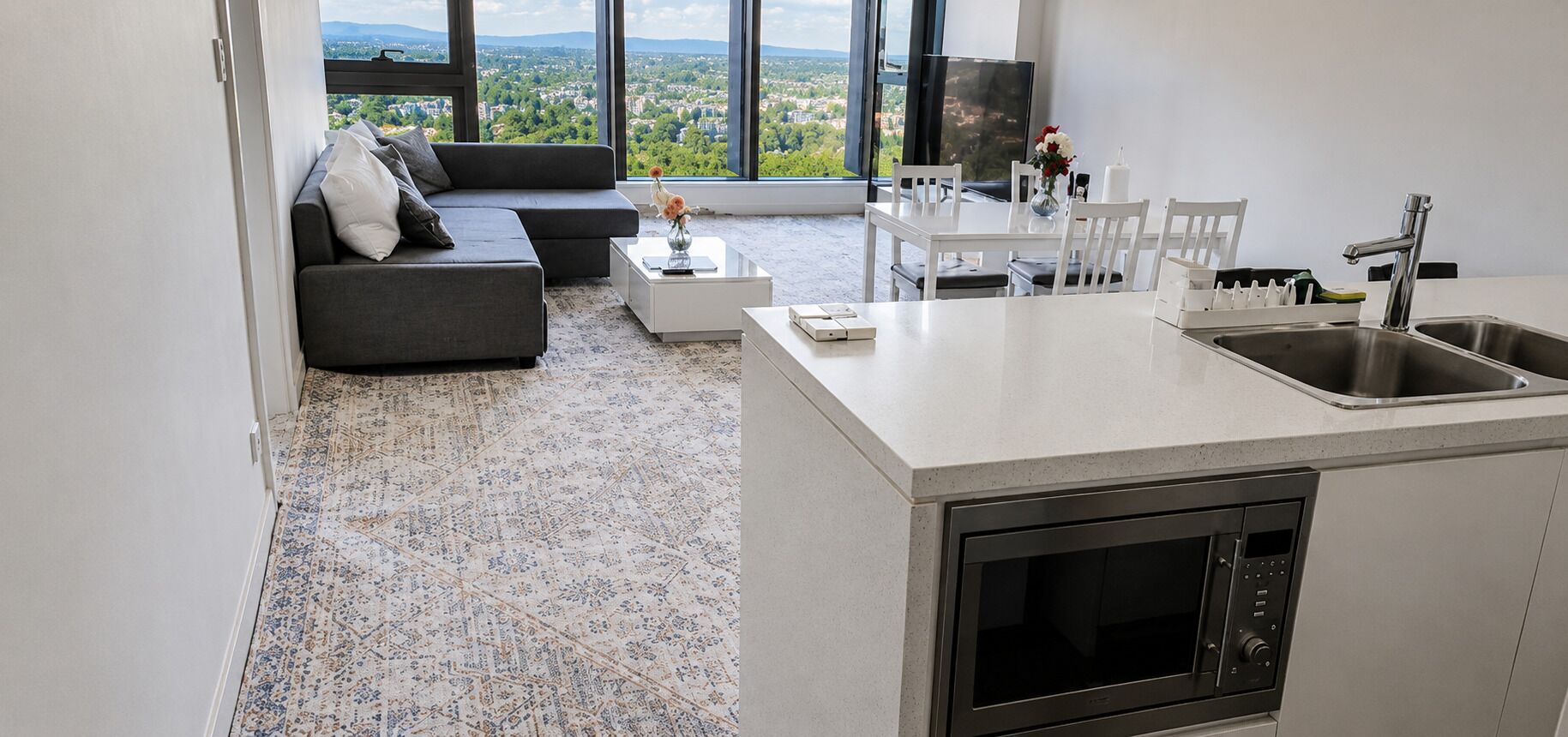 A modern kitchen with a built-in oven, a dining area with a white table and chairs, a grey sofa with cushions, and a large window offering a city view.