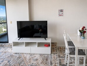 A modern living room with a flat-screen TV on a white entertainment unit, a dining area with a white table and chairs, and a view of the city through a large window. - Cozy apartment Sydney Olympic Park free parking 03 (Sydney Olympic Park)