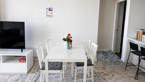 Ein Essbereich mit einem weißen Tisch, vier weißen Stühlen, einer Vase mit Blumen, einem Fernseher auf einem weißen Schrank und einem gemusterten Teppich.