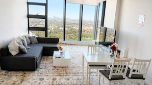 Ein modernes Wohnzimmer mit Sofa, Couchtisch, Esstisch und Stühlen. Große Fenster bieten Blick auf die Stadt.