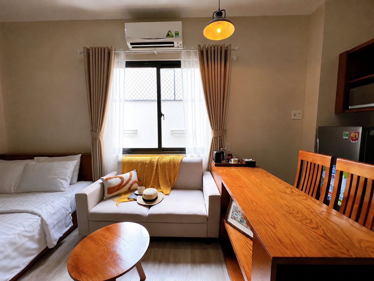 Hotelkamer met een bed, een sofa, een eettafel, een houten bank, een airconditioner, een raam met gordijnen en een hanglamp.