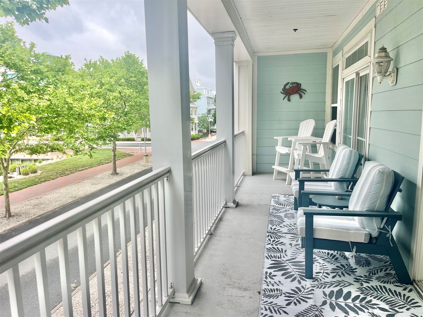 A porch with white railings, a row of white chairs, a patterned rug, and a wall-mounted light fixture.