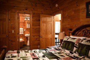 A wooden cabin room with a bed, a nightstand, a lamp, and a bathroom visible through an open door. - Fully Furnished Cabin with Fire Pit Perfect for Small Groups in Oklahoma (Broken Bow)
