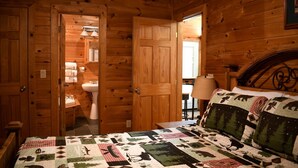 Ein Zimmer einer Holzhütte mit einem Bett, einem Nachttisch, einer Lampe und einem durch eine offene Tür sichtbaren Badezimmer.