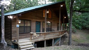 Eine Blockhütte mit Veranda, einer grünen Tür und einem Schild mit einem bogen Baum.