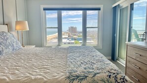 Une chambre avec un grand lit, une table de chevet avec une lampe, une commode et une vue sur la plage et la ville, grâce à une grande fenêtre.
