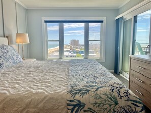 Ein Schlafzimmer mit einem großen Bett, einem Nachttisch mit Lampe, einer Kommode und Blick auf Strand und Stadt durch ein großes Fenster.