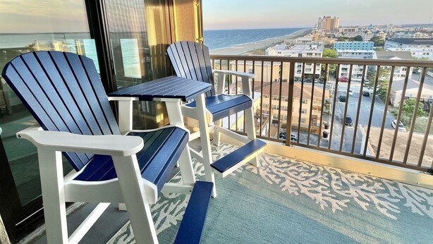 Un balcon avec deux fauteuils Adirondack et une petite table, d’où l’on peut admirer une plage et le paysage urbain.