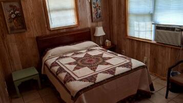 A bedroom with a wooden headboard, a quilted bedspread, a nightstand, a lamp, and a window with blinds.