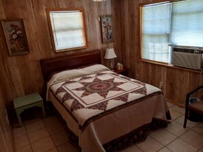 Un dormitorio con cabecera de madera, colcha acolchada, mesita de noche, lámpara y ventana con persianas.