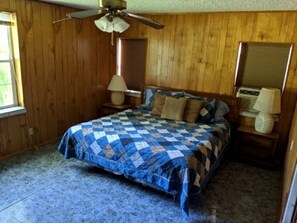 Un dormitorio con cama, ventilador de techo, paredes de madera, una ventana y una mesita de noche con lámpara.
