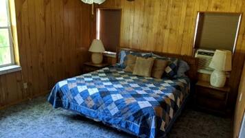 A bedroom with a bed, a ceiling fan, wooden walls, a window, and a nightstand with a lamp.