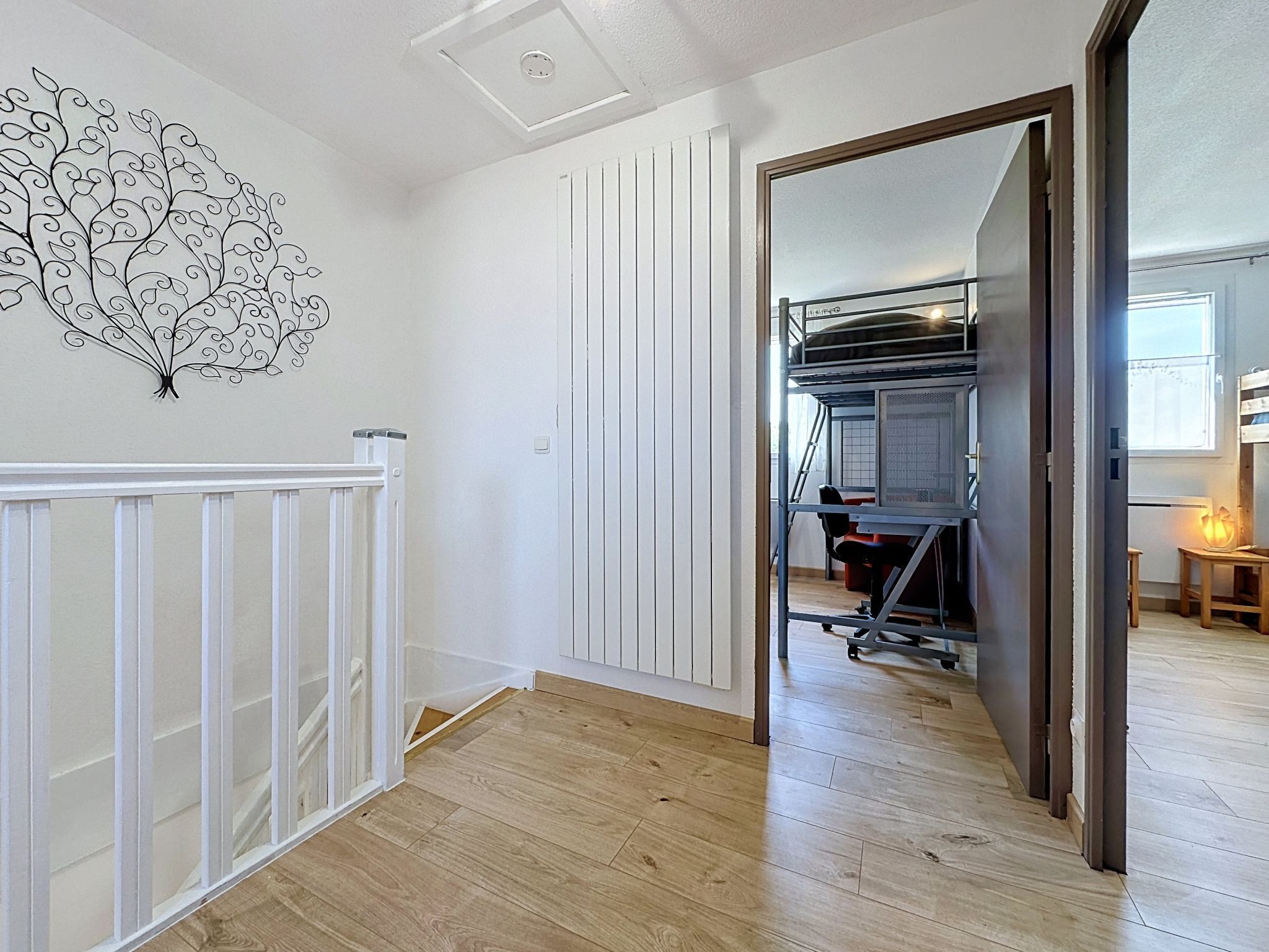 Un pasillo con un radiador blanco, suelo de madera, una puerta blanca y una habitación con una cama litera y un escritorio.
