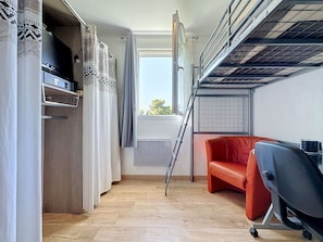 Een kleine kamer met een stapelbed, een rode stoel, een bureau met een computer en een raam met gordijnen.