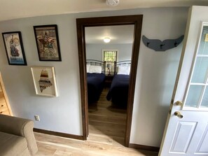 A room with a wooden floor, a door, a bed with a headboard, and wall-mounted artwork. - Cozy Maine Cottage with Outdoor Dining & Modern Comforts near Camden (Lincolnville)