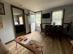 Una stanza con pavimento in legno, un tavolo da pranzo con sedie, una TV fissata al muro e una porta che dà sull'esterno.