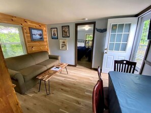 Una habitación con suelo de madera, un sofá, una mesa, una silla y una puerta que da a otra habitación.