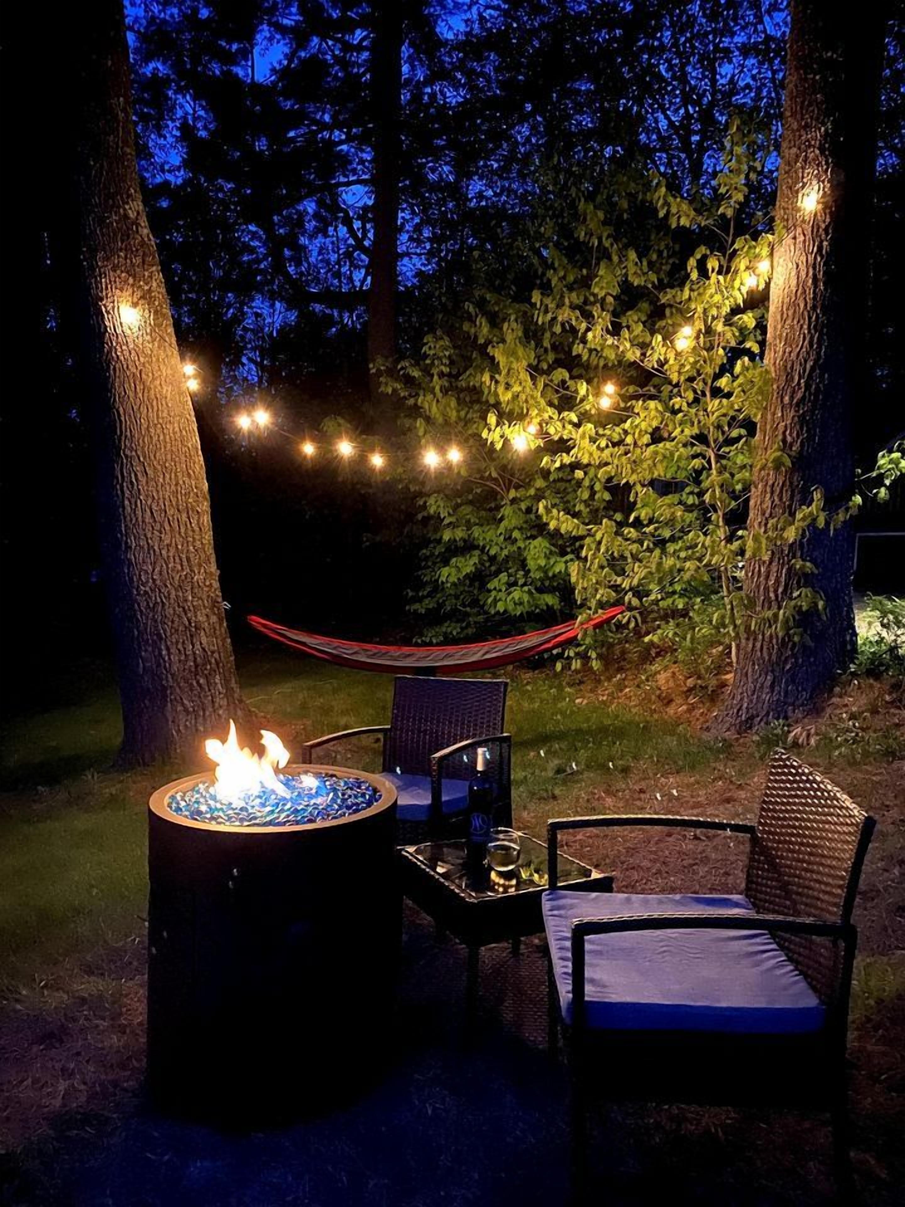 Un espacio al aire libre con asientos nocturnos, una chimenea, dos sillas y luces colgantes.