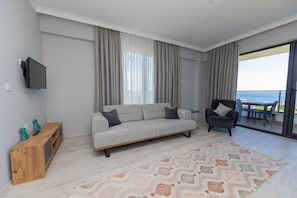 A modern living room with a sofa, a chair, a TV, and a view of the sea through a balcony.
