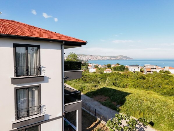 A multi-story building with balconies, a red-tiled roof, and a view of a coastal town and sea. - SİNORA APART (MERKEZ)