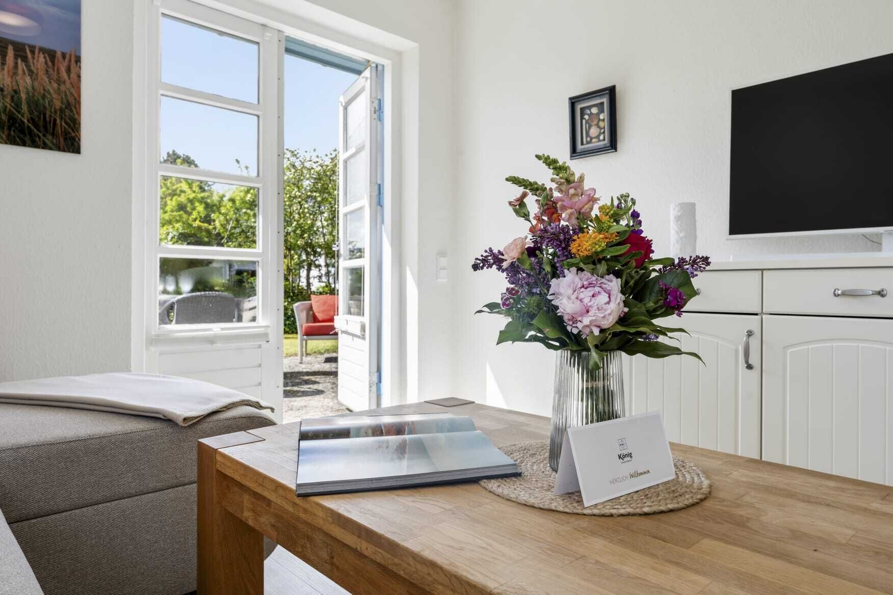 A room with a wooden table, a laptop, a vase with flowers, a framed picture, and a television on the wall.