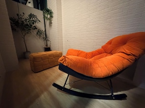 Una habitación con una mecedora, un pequeño reposapiés y dos plantas en macetas frente a una pared de ladrillo blanco.
