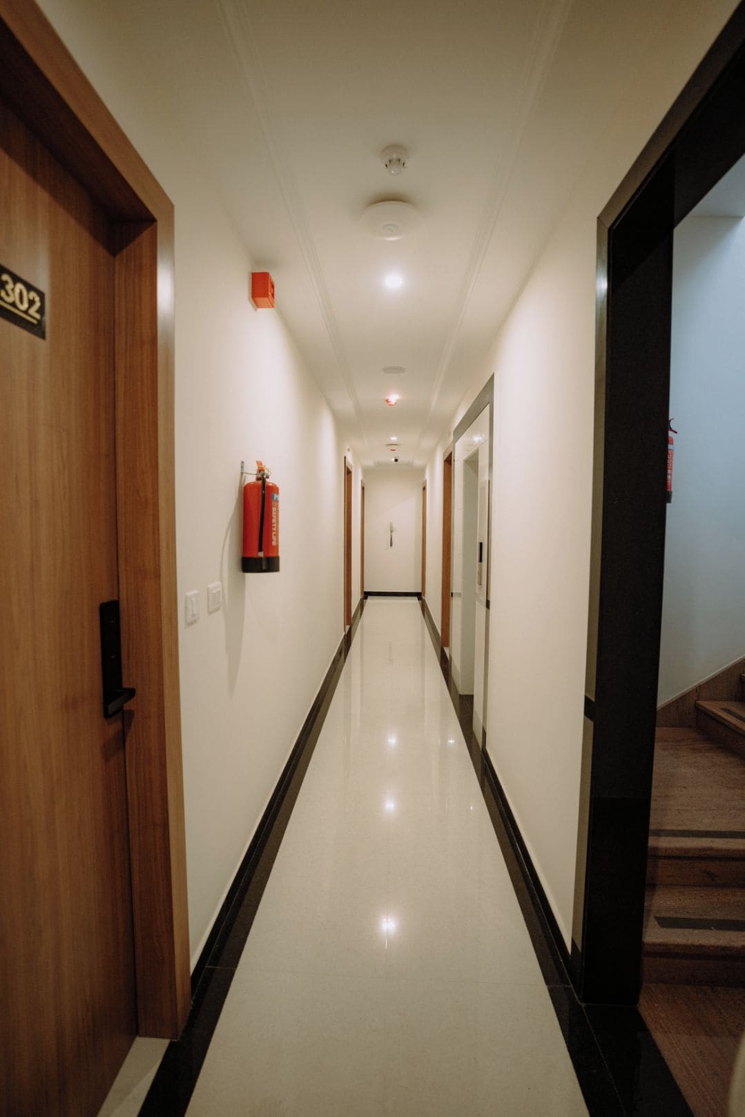 A hotel corridor with a fire extinguisher mounted on the wall, a door with a number sign, and a staircase at the end.