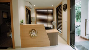 A reception area with a wooden reception desk, a wall-mounted clock, and a window with blinds. - The Camellia Grand  (Lucknow)