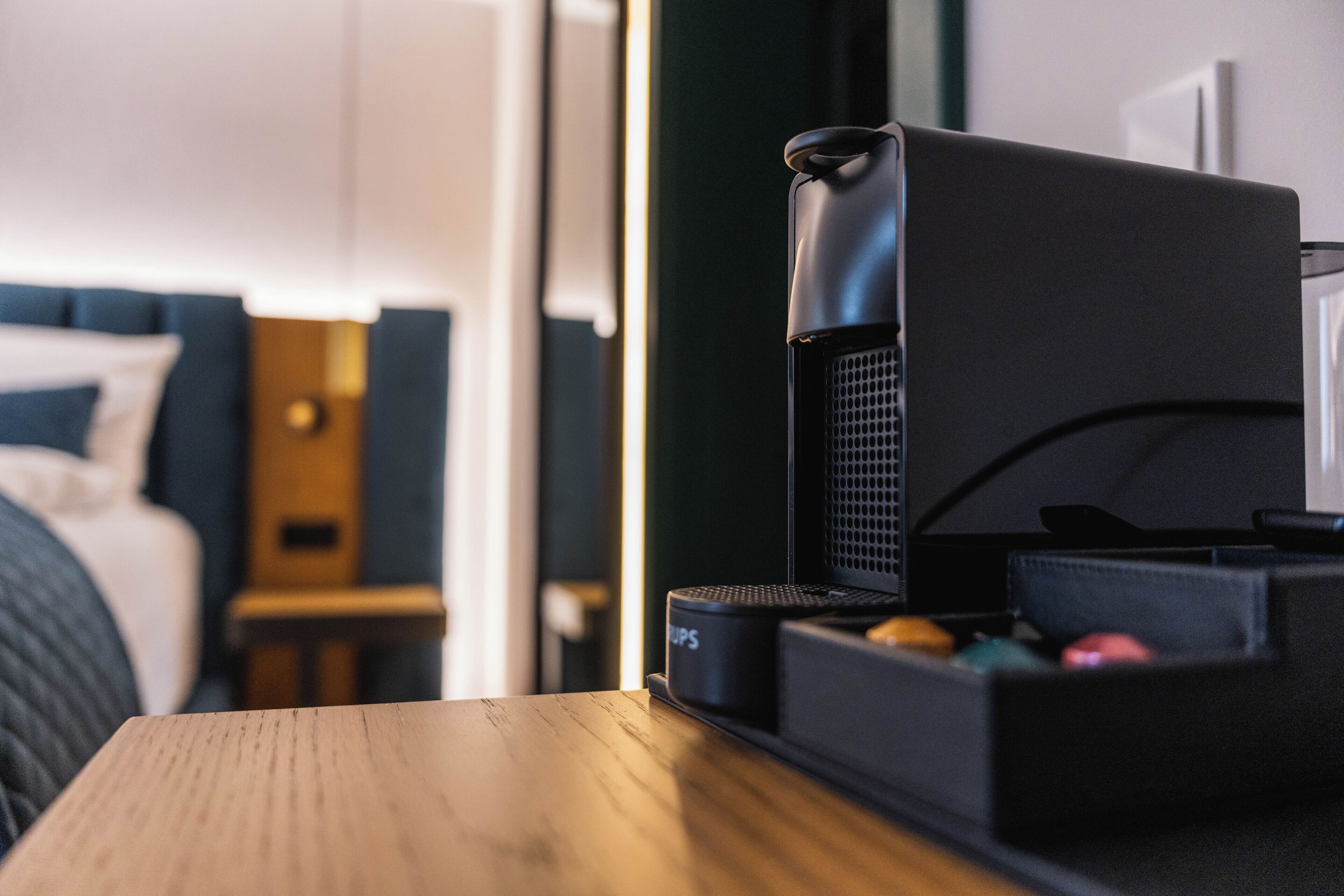 A hotel room with a bed, a wooden nightstand, a black coffee machine, and a black storage box.